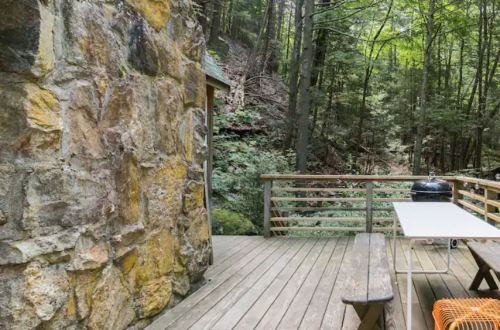 A wooden deck with a stone wall, surrounded by lush green trees and a grill on the side.