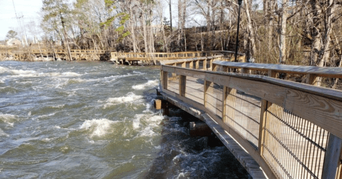 A wooden walkway curves over rushing water, surrounded by trees and a natural landscape.