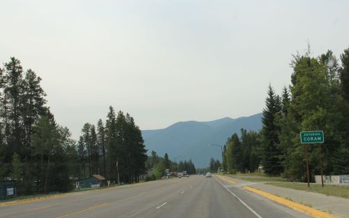 A road leading into Coram, surrounded by trees and mountains under a hazy sky.