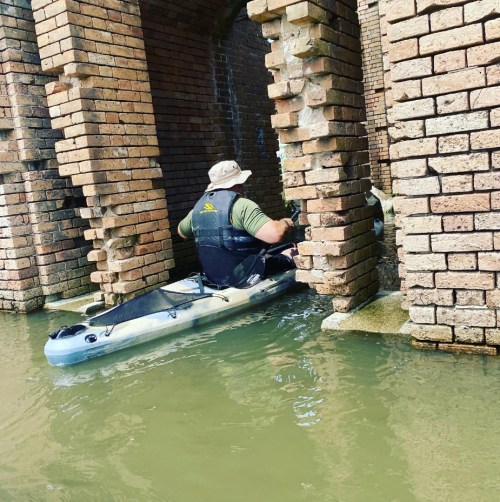 A person in a kayak navigates through brick arches over water, surrounded by a rustic, industrial setting.