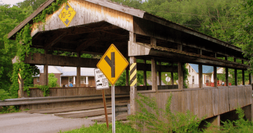 A weathered covered bridge surrounded by greenery, with road signs indicating a speed limit and a curve ahead.
