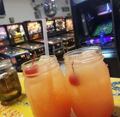 Two cocktails in mason jars with cherries, set against a backdrop of pinball machines.