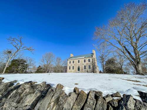 A historic stone house on a snowy landscape, surrounded by bare trees and a rocky wall under a clear blue sky.