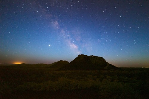 A starry night sky over a rocky landscape, with the Milky Way visible and a distant glow on the horizon.