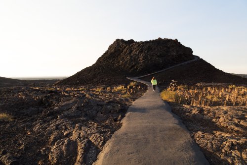 A person walks along a path leading to a rocky hill at sunset, surrounded by barren landscape.