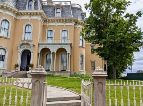 A large, historic mansion with a light-colored facade, ornate details, and a well-maintained lawn and garden.