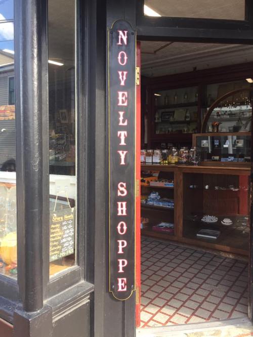 A storefront with a sign reading "Novelty Shoppe," featuring a wooden display inside.