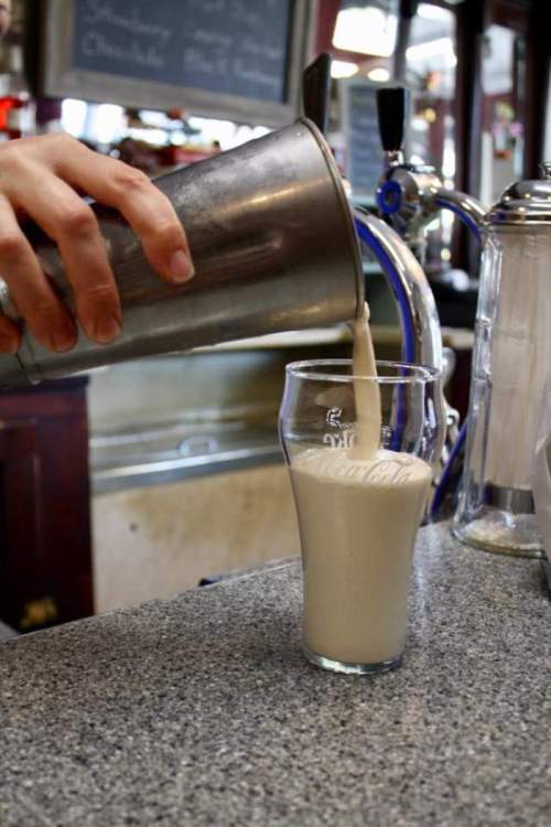 A hand pours a creamy beverage from a metal shaker into a glass on a bar counter.