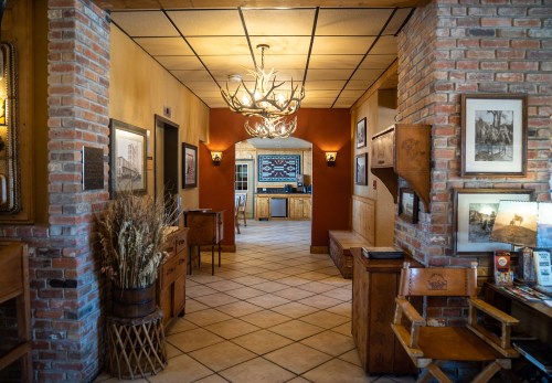 A cozy hallway with brick walls, wooden furniture, and a chandelier, leading to a kitchen area with framed photos.