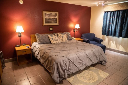 Cozy hotel room with a queen bed, bedside tables, a blue chair, and a window with curtains. Warm lighting creates a welcoming atmosphere.