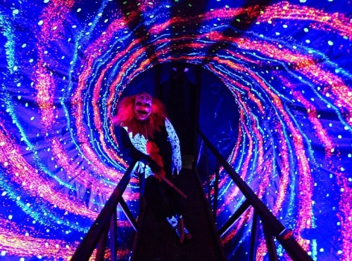 A spooky clown with bright red hair stands in a colorful, swirling tunnel of lights.