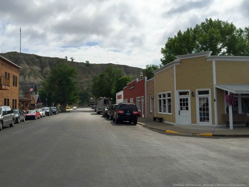 A quiet street in a small town, lined with buildings and parked cars, with a rocky hill in the background under cloudy skies.