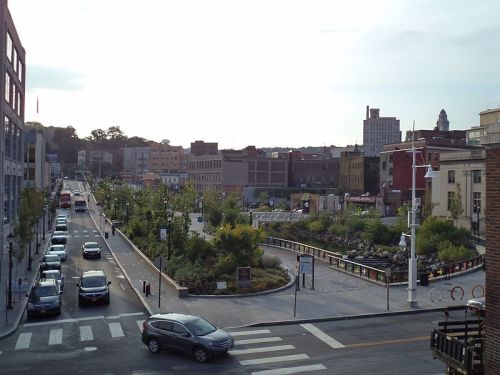 A city street with parked cars, greenery, and buildings under a cloudy sky, showcasing urban landscaping.