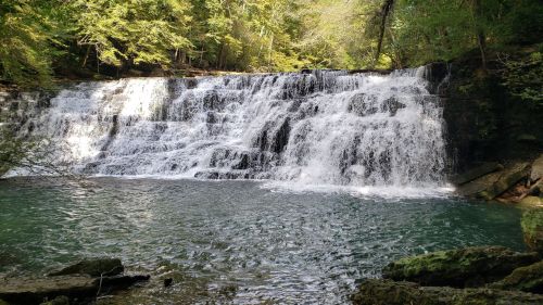 A serene waterfall cascades over rocks into a clear pool, surrounded by lush green trees and sunlight filtering through.
