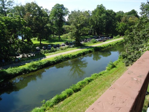 A serene view of a river bordered by lush greenery and trees, with parked cars visible along the bank.