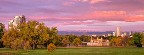 A scenic view of a park at sunset, featuring trees, a lake, and city skyline with mountains in the background.