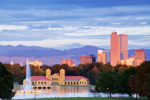 A scenic view of a city skyline with mountains in the background, featuring a lake and a fountain in the foreground.