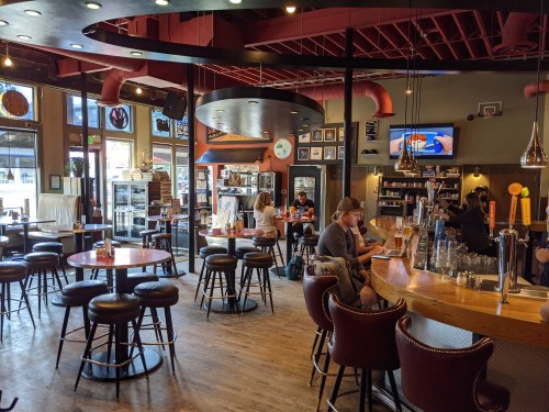 A cozy restaurant interior with wooden tables, bar stools, and patrons enjoying food and drinks.