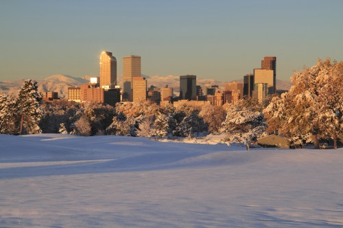 Snow-covered landscape with a city skyline in the background, illuminated by sunlight against a clear blue sky.