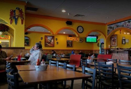 A colorful restaurant interior with tables, chairs, and a TV showing a sports game. Patrons are seated throughout.