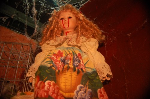 A creepy doll with curly hair and a painted dress, featuring blood on its face, surrounded by cobwebs.