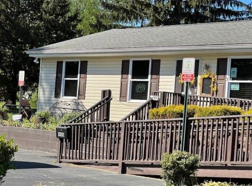 A light-colored house with a sign that says "OPEN," featuring a wooden ramp and landscaped front yard.