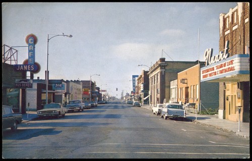 A vintage street scene featuring shops, parked cars, and a bus depot, with a clear sky in the background.