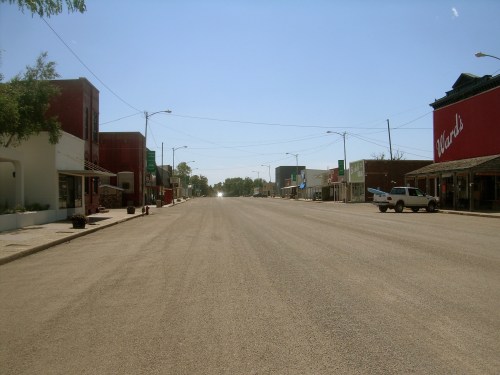 A quiet, empty street lined with small businesses and a clear blue sky.