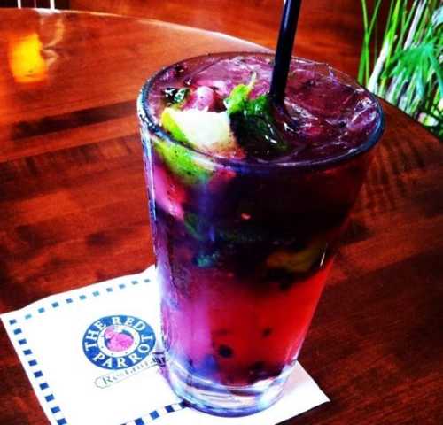 A colorful cocktail with mint, lime, and berries, served in a tall glass on a coaster from The Red Parrot restaurant.