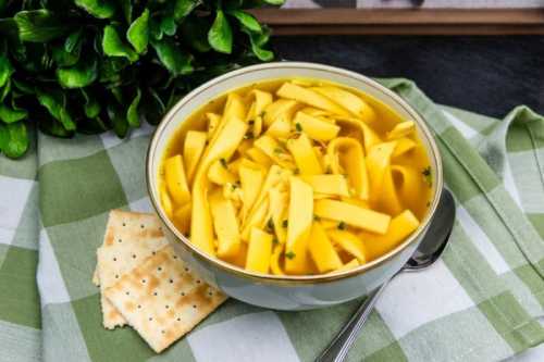 A bowl of noodle soup with yellow noodles, garnished with herbs, next to crackers on a green checkered cloth.