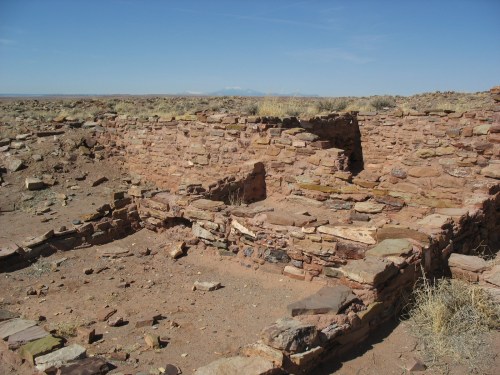 Ruins of an ancient stone structure in a desert landscape, with sparse vegetation and distant mountains under a clear blue sky.