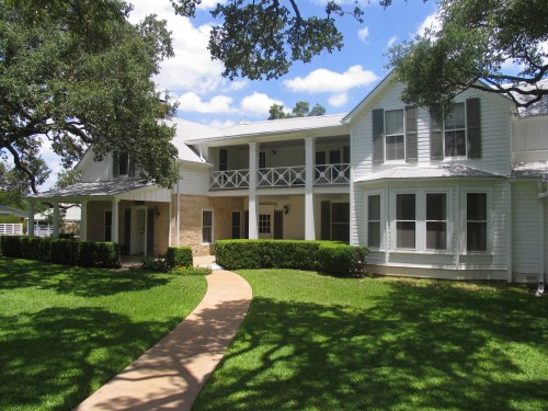 A large, two-story house with a white exterior, surrounded by green grass and trees, under a blue sky with clouds.
