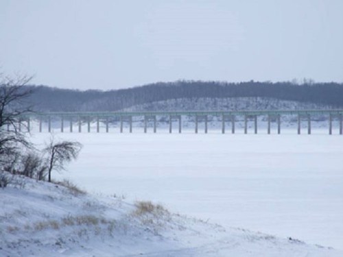 A snowy landscape featuring a bridge spanning a frozen lake, with trees and hills in the background.