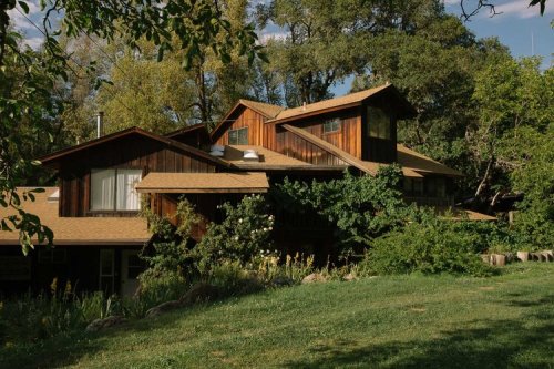 A large, rustic wooden house surrounded by greenery and trees, with multiple roofs and a lush lawn in front.