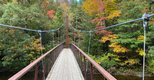 A suspension bridge spans a river, surrounded by vibrant autumn foliage in shades of red, orange, and green.