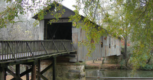 A rustic covered bridge spans a calm river, surrounded by trees and autumn foliage.