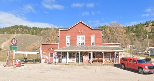 A red wooden building with a porch, surrounded by trees and a blue sky, featuring a gas station sign and parked red truck.