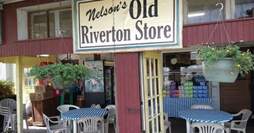 A rustic storefront with a sign reading "Nelson's Old Riverton Store," featuring outdoor seating and hanging plants.