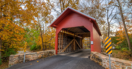 A red covered bridge surrounded by autumn foliage and a stone entrance, with a paved road leading through it.
