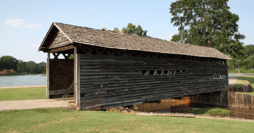A rustic wooden covered bridge over a small stream, surrounded by green grass and trees under a clear blue sky.