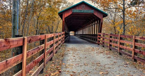 A wooden covered bridge surrounded by autumn trees and fallen leaves, leading into a bright, open pathway.