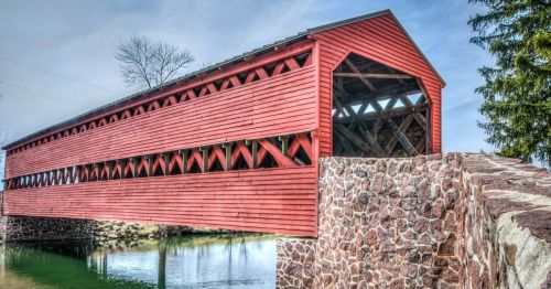 A red covered bridge spans a calm waterway, surrounded by trees and a stone support structure.