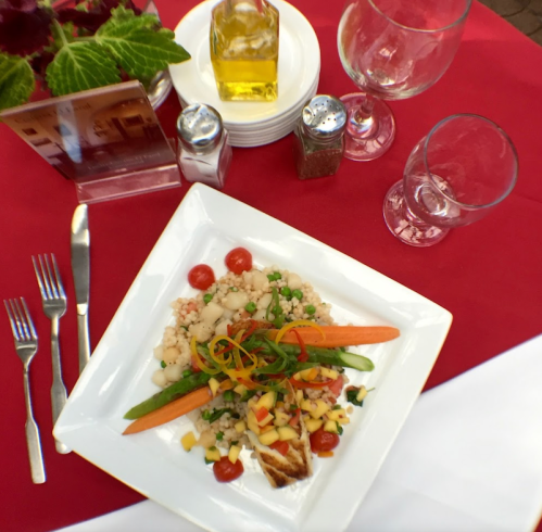 A plated meal featuring grilled fish, colorful vegetables, and grains, set on a red tablecloth with dining utensils.