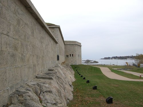 A stone fortification along a grassy path, overlooking a calm body of water under a cloudy sky.