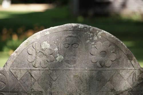 A weathered gravestone with floral carvings and a face-like engraving, set against a blurred green background.
