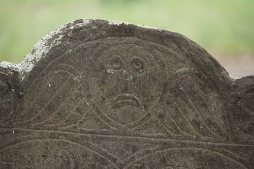 Close-up of a weathered gravestone featuring a carved face with a surprised expression and two wings.