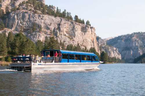 A blue tour boat with passengers glides through a calm lake, surrounded by rocky cliffs and lush trees.