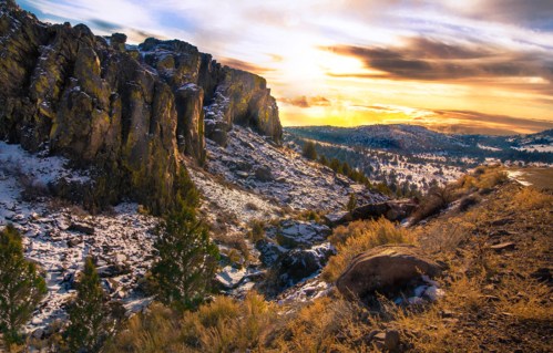 A scenic landscape featuring rocky cliffs, snow-covered ground, and a vibrant sunset sky.