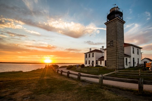 A lighthouse stands by the water at sunset, with colorful clouds and a grassy area in the foreground.