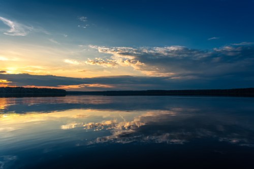 A serene lake at sunset, reflecting colorful clouds and a blue sky, surrounded by trees on the horizon.